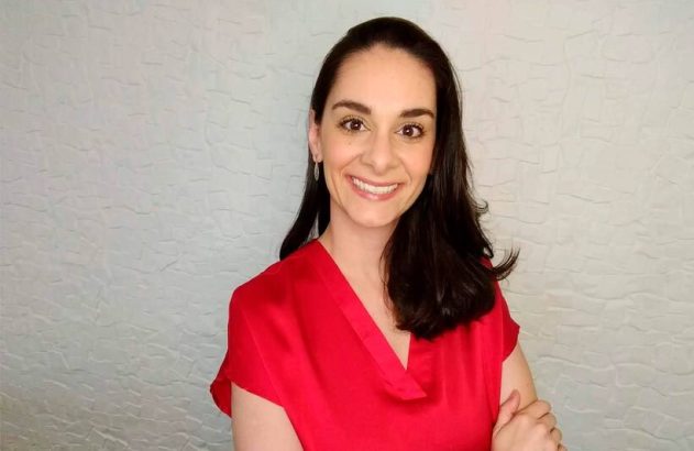 Mulher sorrindo de blusa vermelha e cabelo escuro, braços cruzados, em frente a parede texturizada.