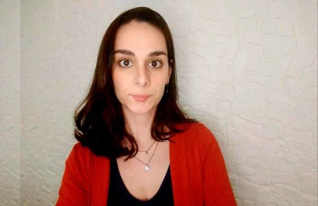 Mulher de cabelo escuro e casaco laranja olha para a câmera. Fundo de parede branca texturizada.