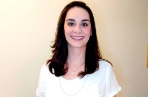 Mulher sorridente de cabelos escuros em camiseta branca, olhando para frente.