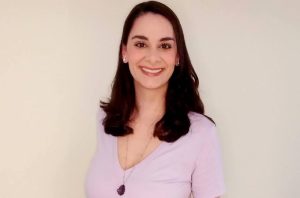 Retrato de mulher sorrindo, cabelos escuros, blusa lavanda e colar. Fundo neutro.