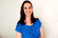 Mulher de cabelo escuro sorrindo, vestindo blusa azul contra parede branca.