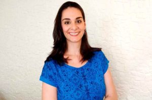 Mulher de cabelo escuro sorrindo, vestindo blusa azul contra parede branca.