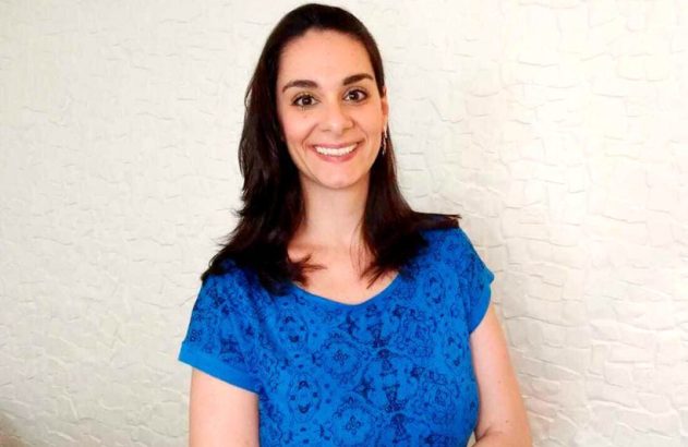 Mulher de cabelo escuro sorrindo, vestindo blusa azul contra parede branca.