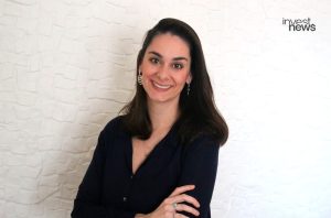 Mulher sorridente com cabelo escuro, camisa azul e braços cruzados. Fundo branco com logo "invest news".