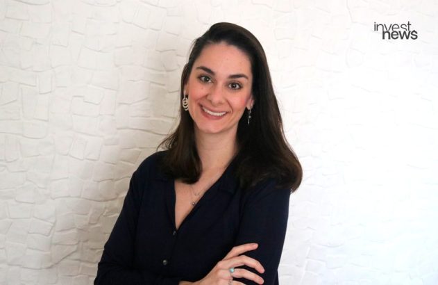 Mulher sorridente com cabelo escuro, camisa azul e braços cruzados. Fundo branco com logo "invest news".