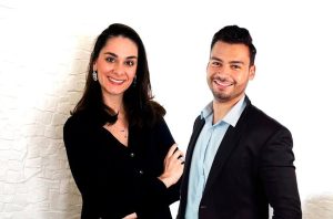 Homem e mulher sorrindo para a câmera, em trajes profissionais, fundo branco.