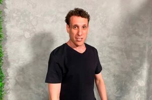 Homem de camiseta preta e cabelo cacheado sorri. Fundo cinza com textura e planta verde.