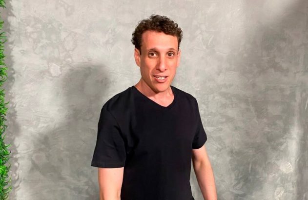 Homem de camiseta preta e cabelo cacheado sorri. Fundo cinza com textura e planta verde.