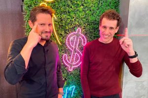 Dois homens sorrindo, um com o dedo na têmpora e outro apontando para cima, em frente a um neon de cifrão em uma parede de folhagem.