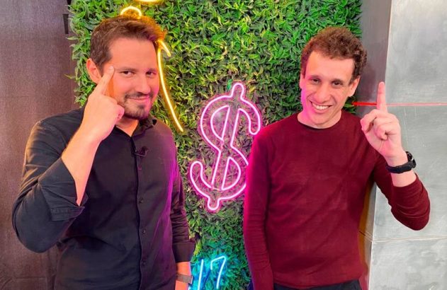 Dois homens sorrindo, um com o dedo na têmpora e outro apontando para cima, em frente a um neon de cifrão em uma parede de folhagem.