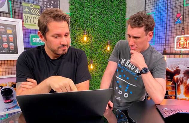 Dois homens sentados a uma mesa olham um laptop. Um toca a tela, o outro observa atentamente.