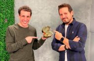 Dois homens sorrindo apontam para um sinal de dólar dourado que um deles segura.