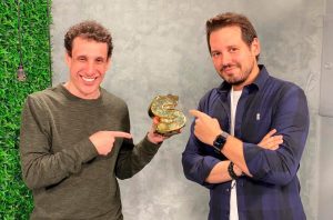 Dois homens sorrindo apontam para um sinal de dólar dourado que um deles segura.