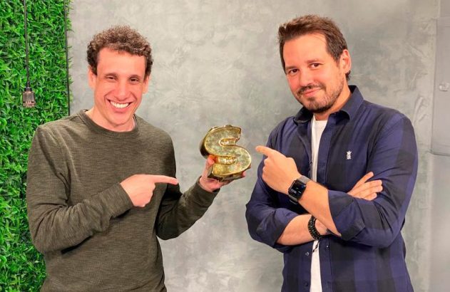 Dois homens sorrindo apontam para um sinal de dólar dourado que um deles segura.