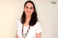 Mulher sorrindo, com cabelos escuros, camiseta branca e colar de contas coloridas.