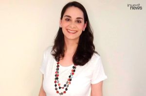 Mulher sorrindo, com cabelos escuros, camiseta branca e colar de contas coloridas.