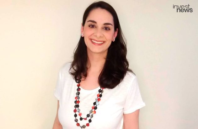 Mulher sorrindo, com cabelos escuros, camiseta branca e colar de contas coloridas.