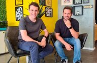 Dois homens sorrindo, sentados em cadeiras, em um escritório com paredes amarelas e cinzas.