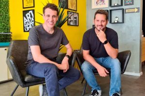 Dois homens sorrindo, sentados em cadeiras, em um escritório com paredes amarelas e cinzas.