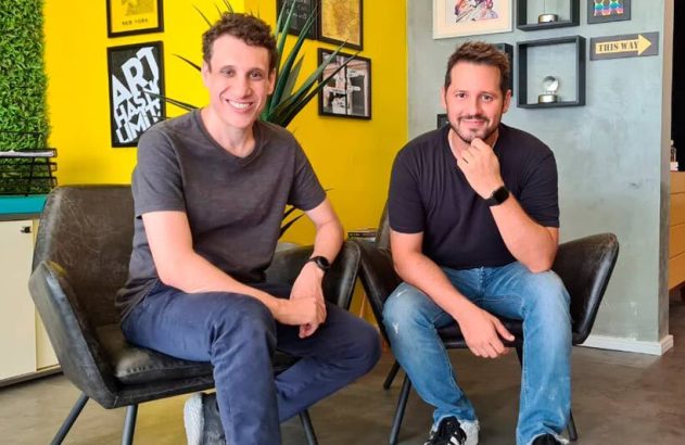 Dois homens sorrindo, sentados em cadeiras, em um escritório com paredes amarelas e cinzas.