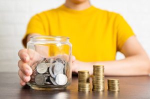 Pessoa economizando: mão segura pote de moedas prateadas; pilhas de moedas douradas na mesa.