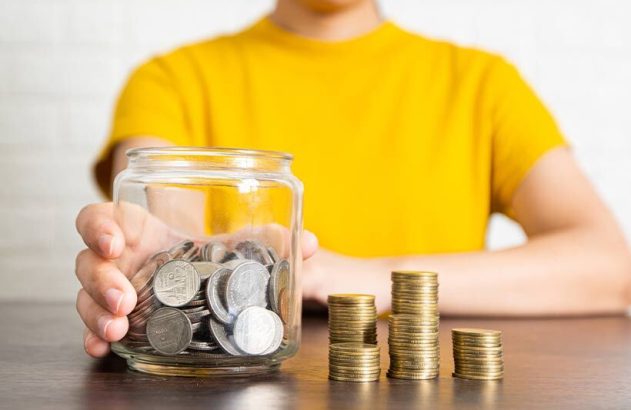 Pessoa economizando: mão segura pote de moedas prateadas; pilhas de moedas douradas na mesa.