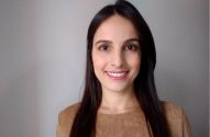 Mulher sorrindo, cabelo escuro, blusa marrom, olhando para a câmera. Fundo cinza claro.