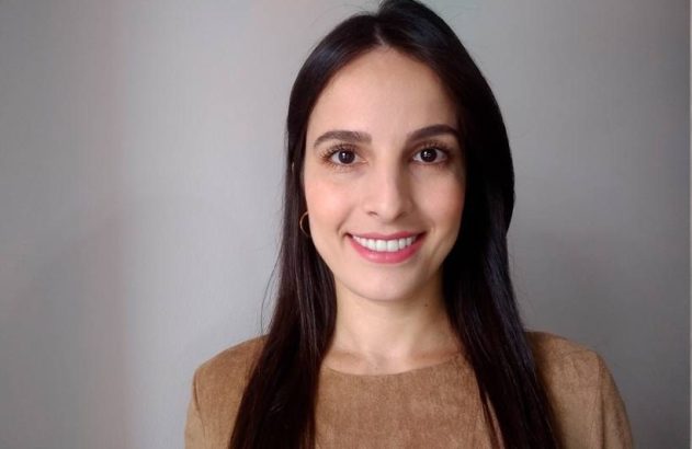 Mulher sorrindo, cabelo escuro, blusa marrom, olhando para a câmera. Fundo cinza claro.