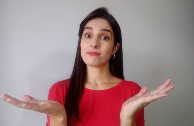 Mulher de cabelos escuros e camisa vermelha, com expressão de dúvida e mãos abertas, palmas para cima.