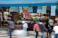 Feira de rua com barraca de roupas. Pessoas borradas por movimento e manequins exibem vestuário sob lona azul.
