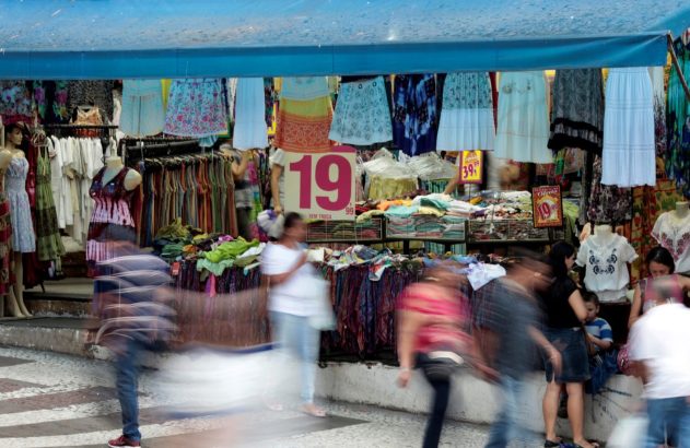 Feira de rua com barraca de roupas. Pessoas borradas por movimento e manequins exibem vestuário sob lona azul.