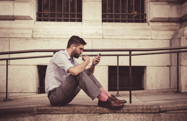 Um homem sentado ao ar livre, olhando o celular, com fones de ouvido e usando um vape.