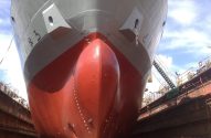 Navio em dique seco, visto da proa. Casco vermelho e cinza, estruturas do dique e céu azul. Em manutenção ou reparo.