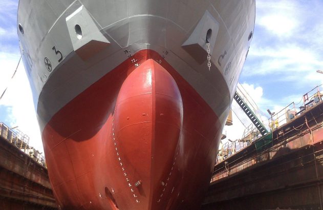 Navio em dique seco, visto da proa. Casco vermelho e cinza, estruturas do dique e céu azul. Em manutenção ou reparo.