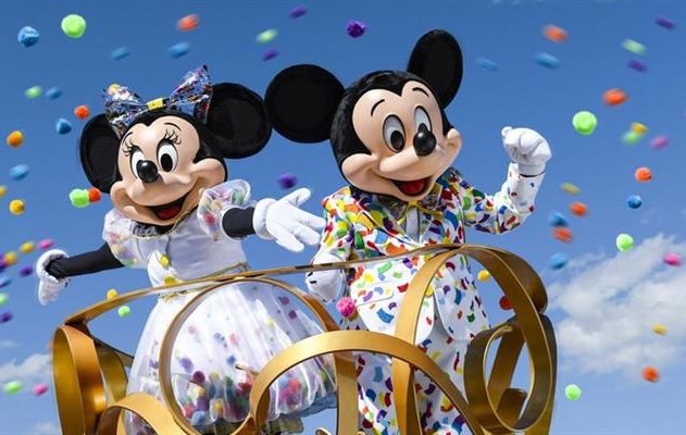 Mickey e Minnie Mouse em desfile, com confetes coloridos caindo. Ambos em trajes brancos estampados, sob céu azul.