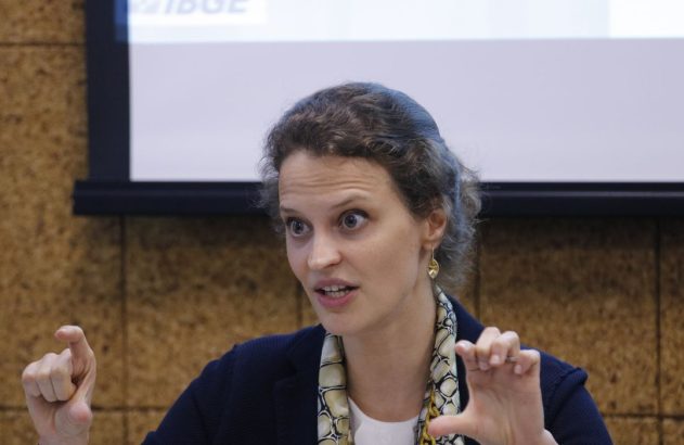 Mulher de olhos arregalados gesticula com as mãos, indicando algo pequeno. Usa lenço estampado; telão ao fundo.