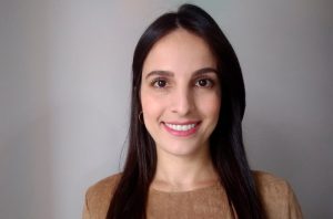 Mulher de cabelo escuro sorrindo e olhando para a câmera. Fundo cinza.