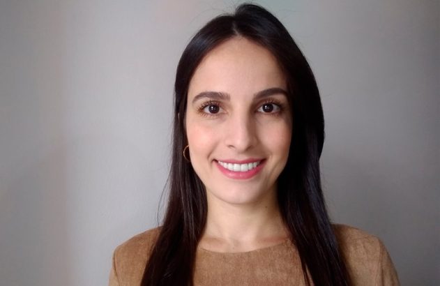 Mulher de cabelo escuro sorrindo e olhando para a câmera. Fundo cinza.