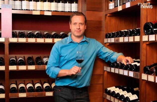 Homem em adega, camisa azul, segurando taça de vinho tinto. Mão em garrafas nas prateleiras.