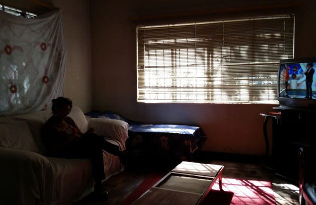 Mulher sentada em sala escura com sol forte vindo da janela. Uma TV está ligada à direita.