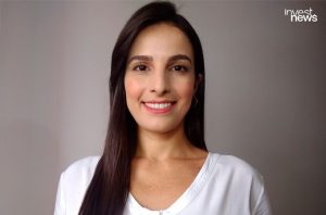 Mulher jovem com cabelo escuro e liso, sorrindo e olhando para a câmera, vestindo blusa branca.