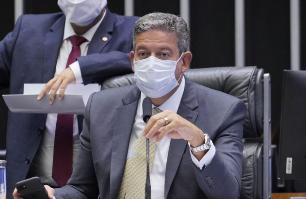 Parlamentar de terno e máscara sentado em plenário, com microfone. Outro homem de terno e máscara em pé ao fundo.