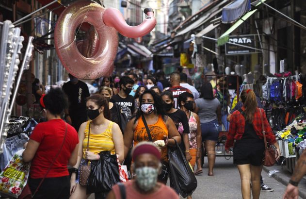 Rua movimentada com muitas pessoas, algumas de máscara, passando por lojas com um inflável rosa de flamingo pendurado.