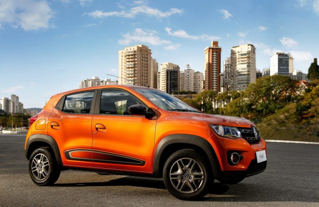 Renault Kwid laranja estacionado em asfalto, com prédios e céu azul no fundo.