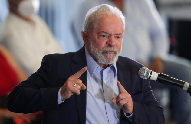 Homem de barba e cabelo grisalhos, de terno, gesticula com dedos indicadores ao falar no microfone.