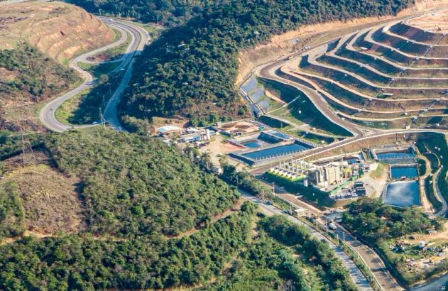 Paisagem aérea: grande aterro/mina em socalcos, estrada, instalações industriais e florestas.