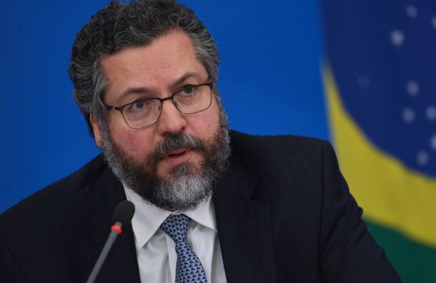 Homem barbudo de óculos e terno fala ao microfone, com bandeira do Brasil ao fundo.