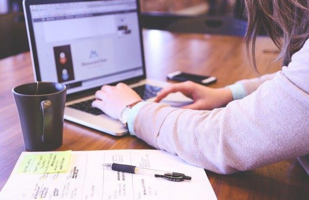 Pessoa trabalhando em laptop sobre mesa com xícara de café e anotações.