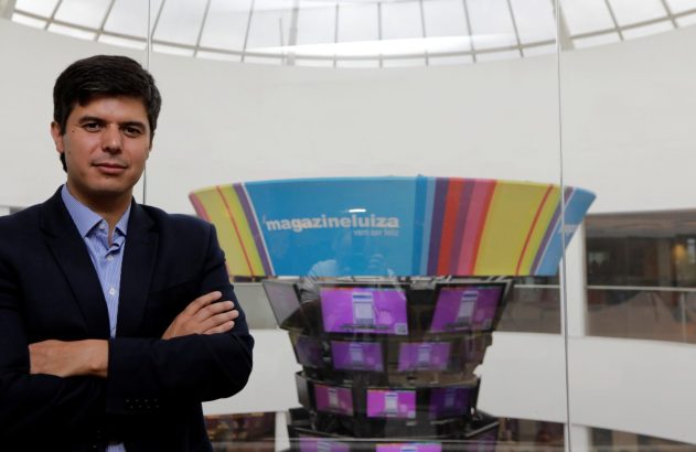 Homem de terno, braços cruzados, posa em ambiente de escritório com display da Magazine Luiza ao fundo.