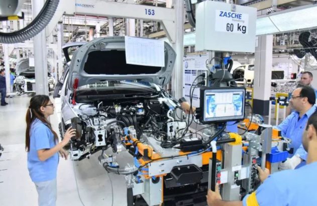 Trabalhadores montam veículo em fábrica. Homem opera equipamento com monitor, mulher inspeciona carro incompleto.